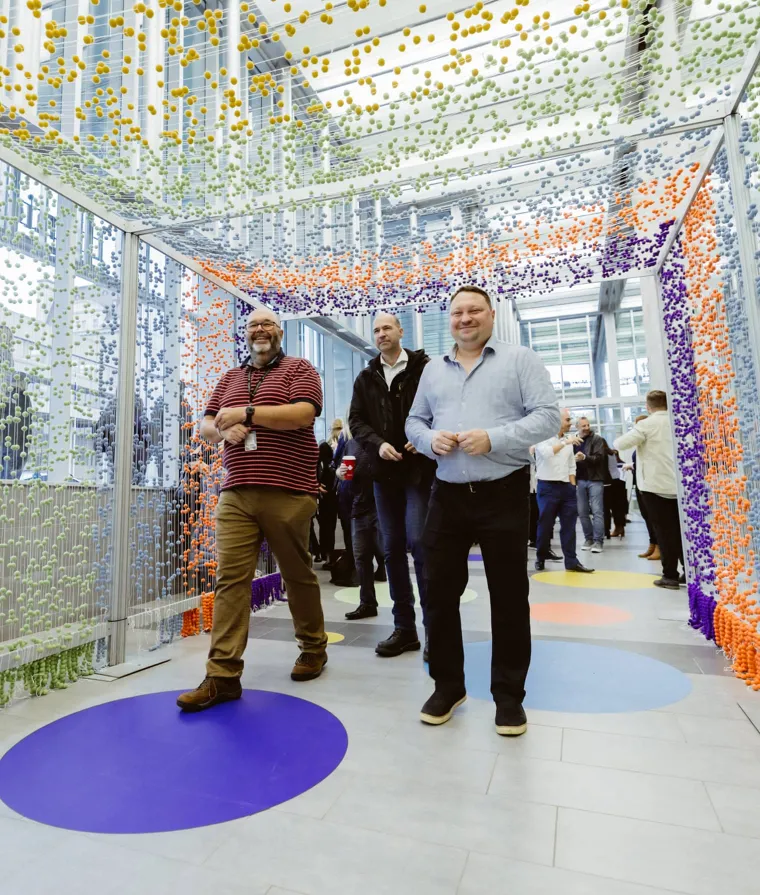 Personnes sous une installation artistique colorée avec d’autres personnes en arrière‑plan